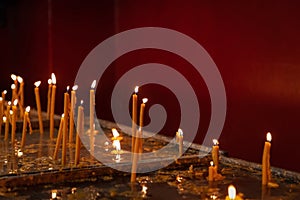 Lit thin wax candles standing in candleholders on church table against a red wall,Â religion concept